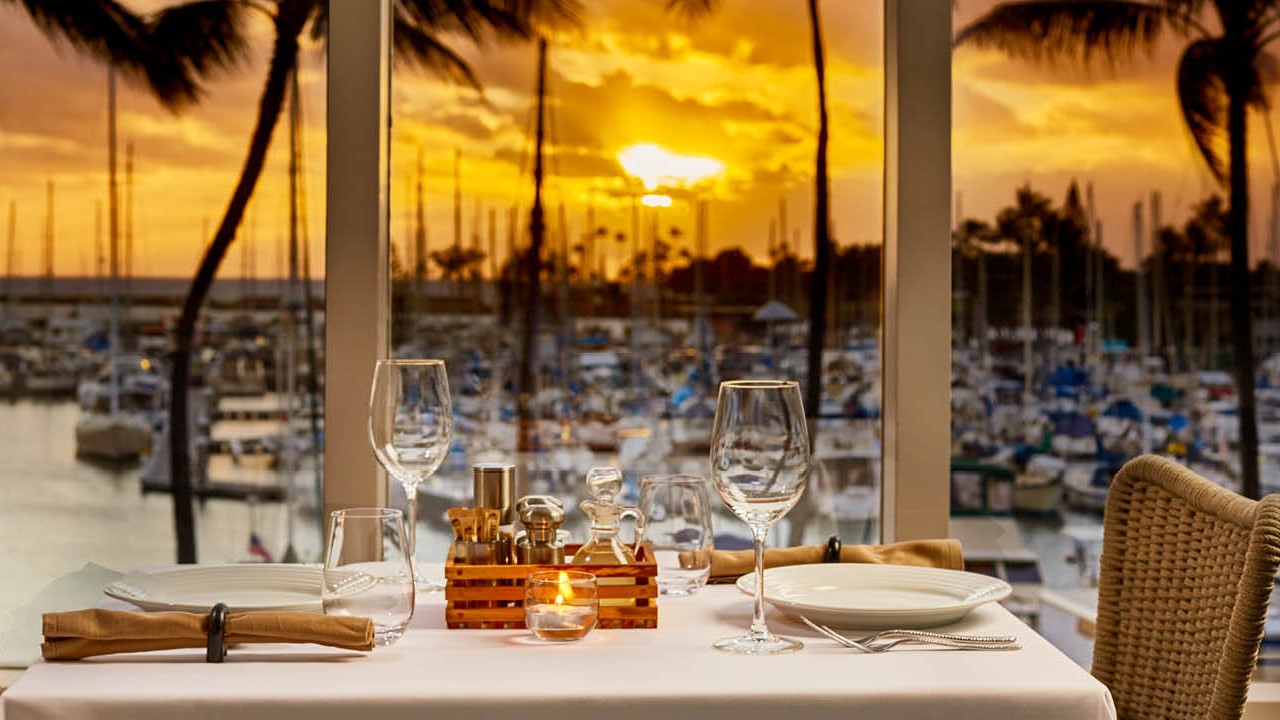A set table at 100 Sails at sunset, with the yacht masts of Ala Wai Harbor and a gold sky through the window