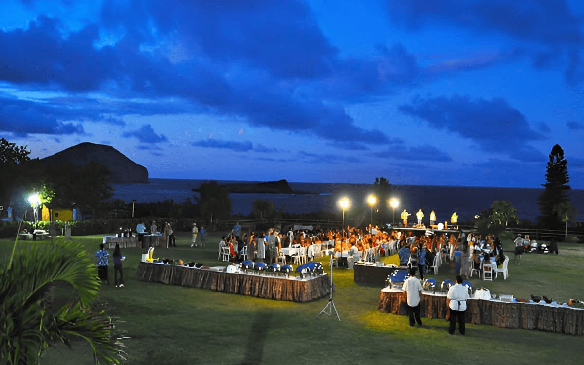 The lawn at Sea Life Park at dusk, guests at long tables beside the buffet with torches lit and Manana Island offshore