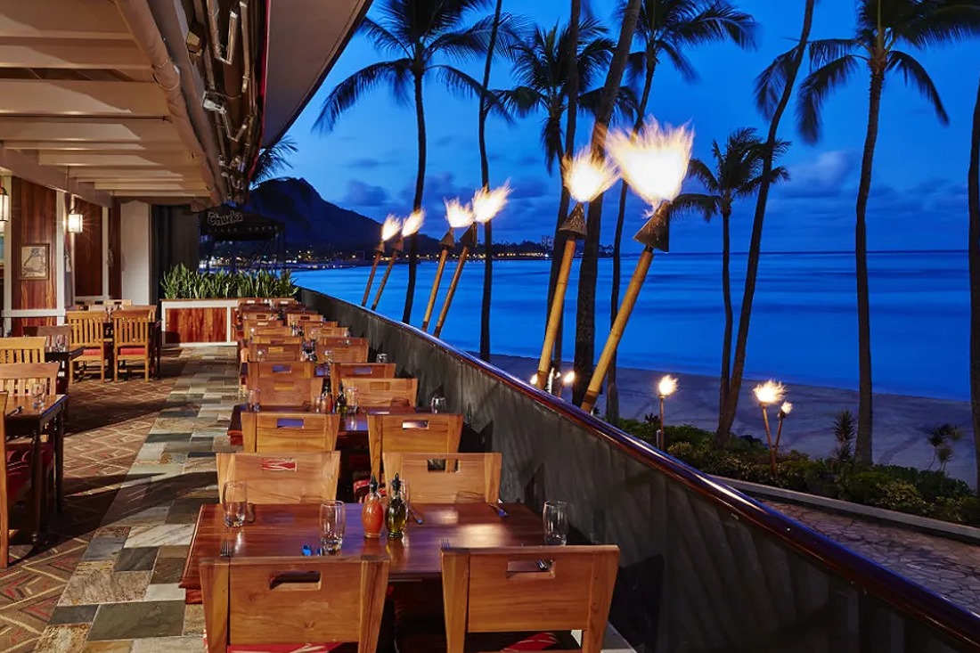 The open-air lanai at Hula Grill Waikiki at dusk, with tiki torches, palms and Diamond Head beyond the beach