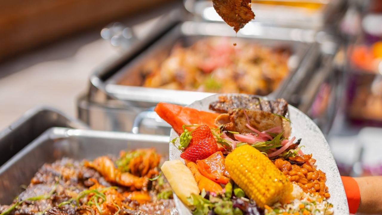 A plate of Hawaiian barbecue at Queensbreak with grilled meat, corn on the cob, baked beans and fruit, in front of the buffet trays