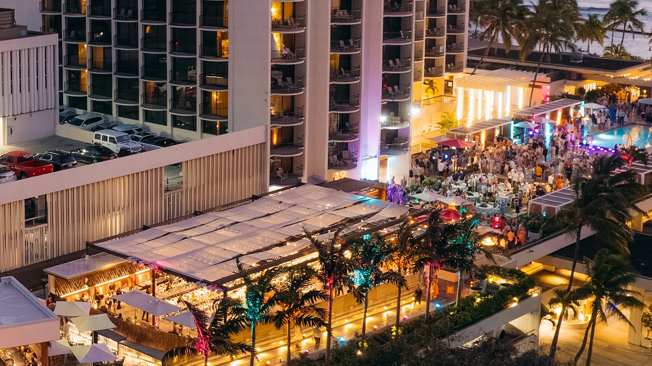 The lit open-air deck at Queensbreak in the evening, with tables under the canopy, palms and the pool crowd beside Waikiki Beach